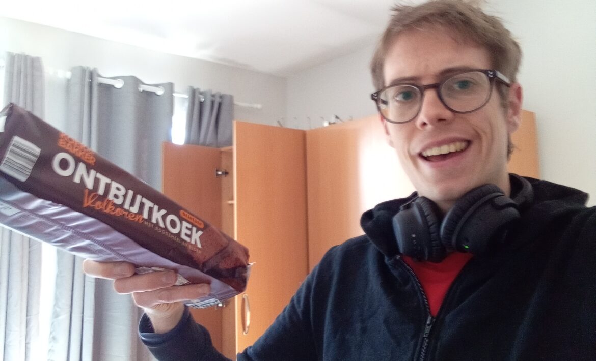A white man (Jorik) wearing glasses and headphones around his neck. He is also wearing a black hoodie and red t-shirt. He is holding up a packet of Ontbijtkoek and wears a quizzical expression.