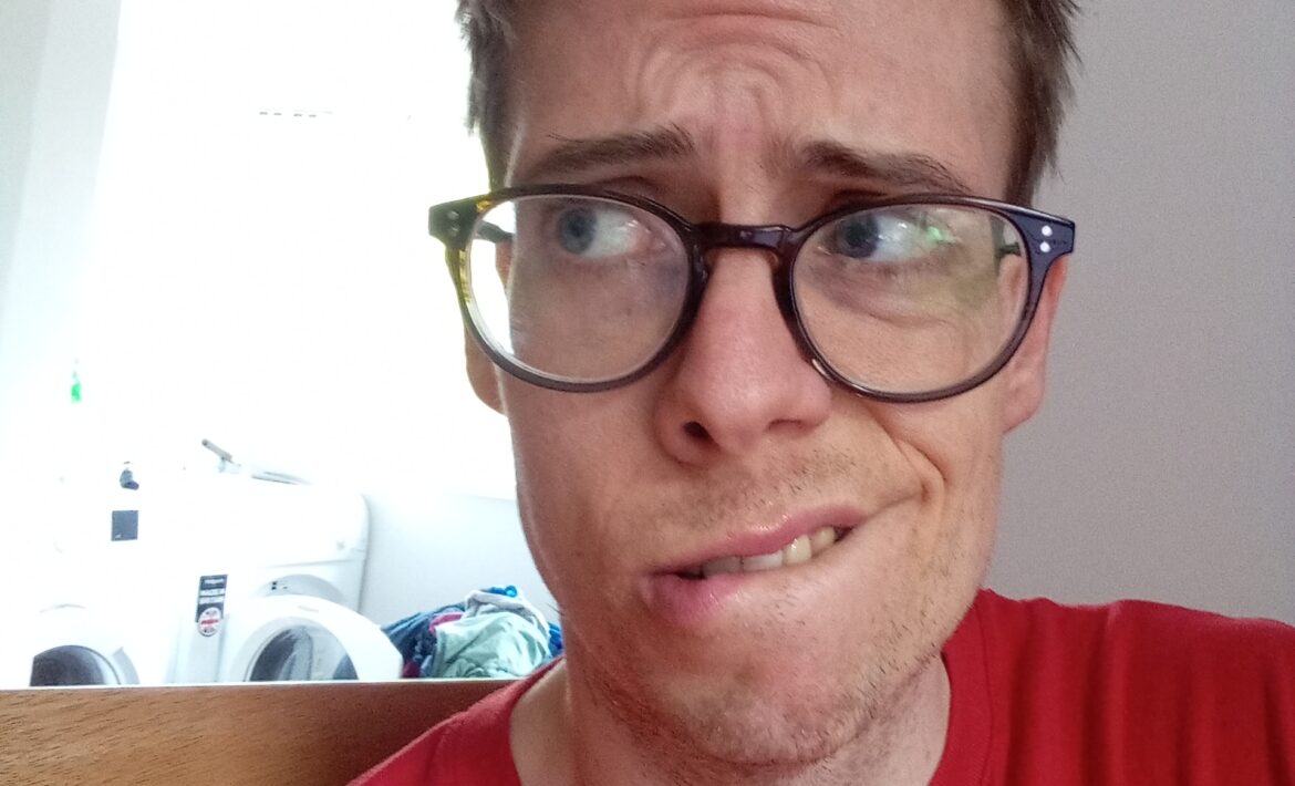 A picture of Jorik, a bespectacled autistic gay man in his thirties who hasn't had a shave for a few days. He has short, dark blond hair and is wearing a red Outdoorlads t-shirt. His face looks worried, biting his lip.