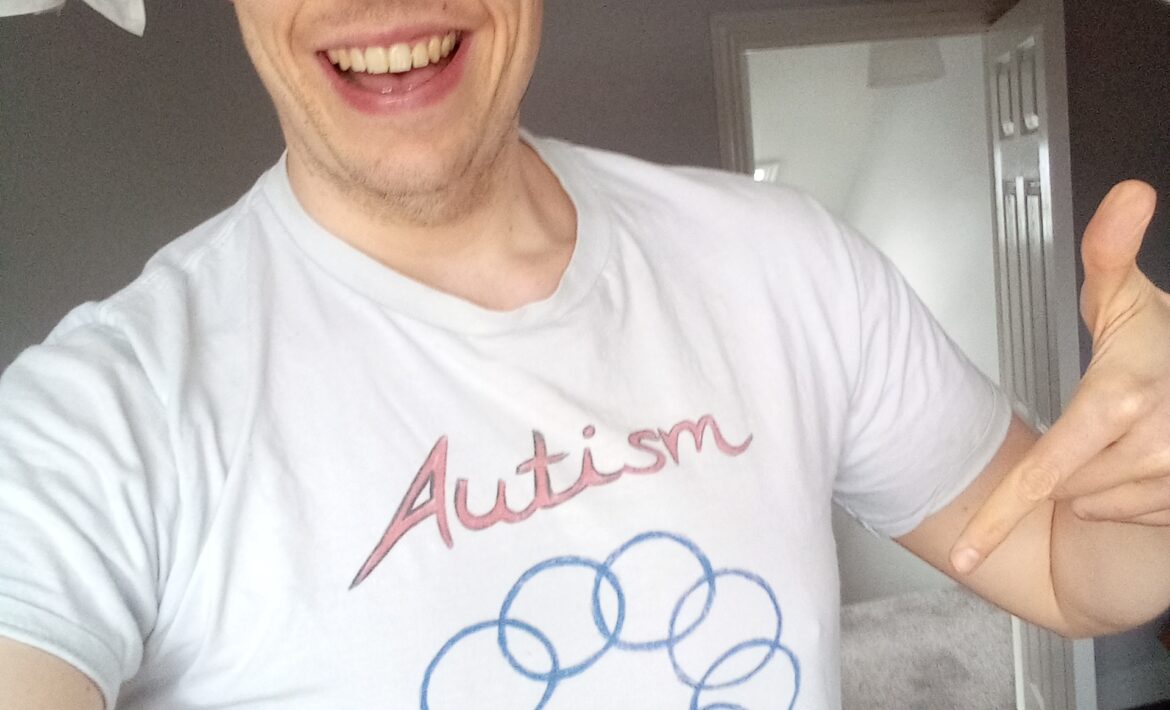 A 33 year old white autistic with light blonde hair is pointing at his white t-shirt, which says "Autism" and Embrace Diversity". Between the two words is an image of a series of interlocking circles, culminating in a heart with rainbow colours.