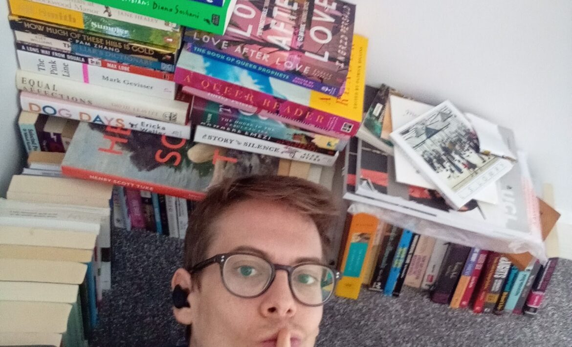 Jorik (a thirty-three year old wearing glasses, a faded blue shirt and wireless earbuds) is lying down next to a handsome stack of books, all queer, while pursing his lips and holding his finger in a 'shhh' motion.