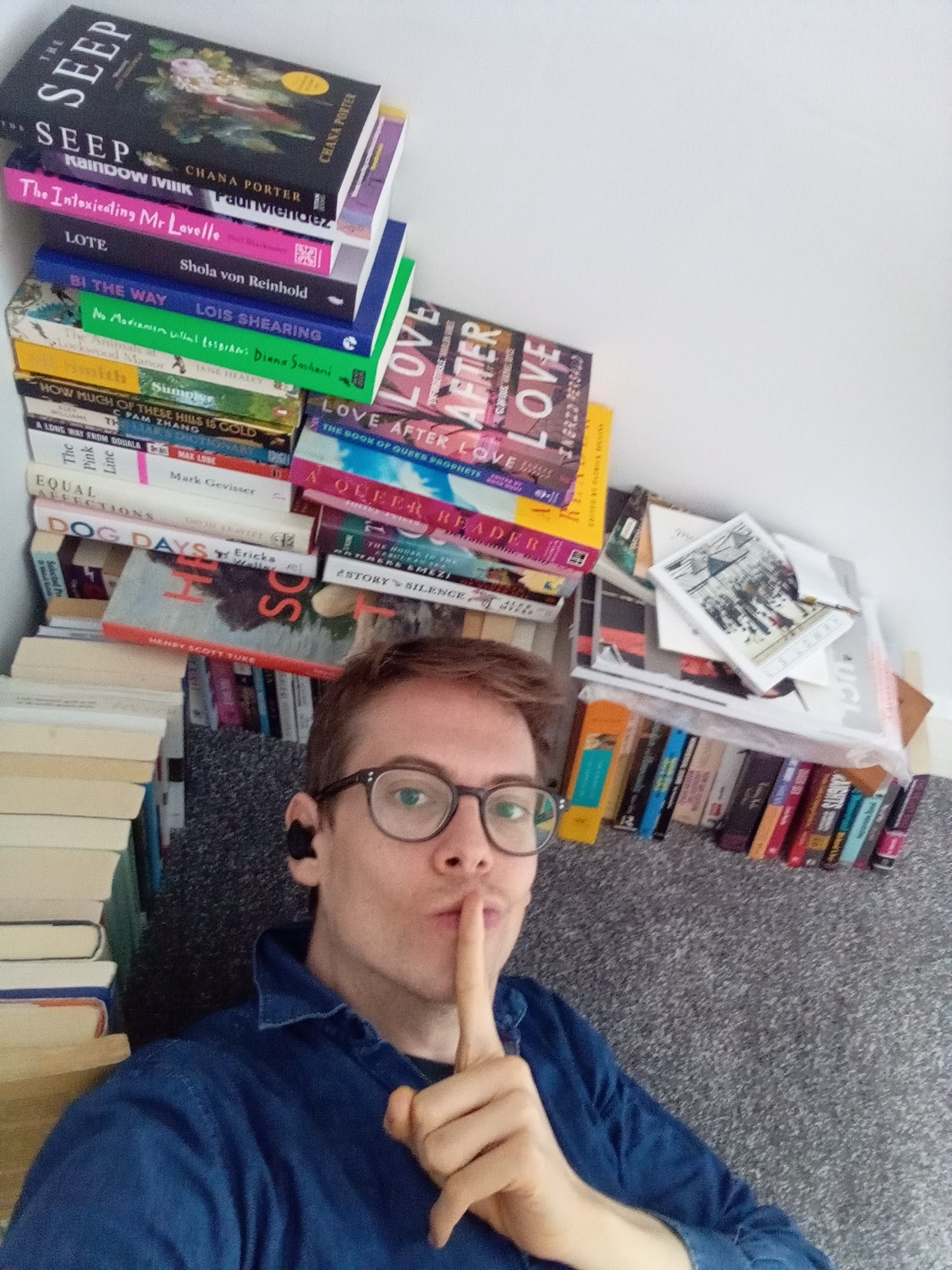Jorik (a thirty-three year old wearing glasses, a faded blue shirt and wireless earbuds) is lying down next to a handsome stack of books, all queer, while pursing his lips and holding his finger in a 'shhh' motion.
