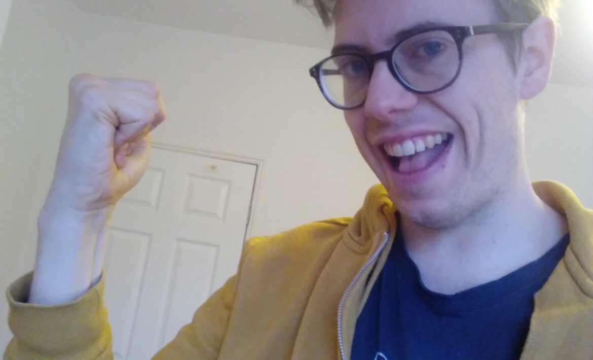 Pictured: a white autistic manchild of nearly 34, wearing glasses, fluffy hair, an old University of Amsterdam t-shirt and a yellow hoodie. Jorik is holding his fist up in a power-stance, while smiling broadly, teeth showing smugly. I love him.