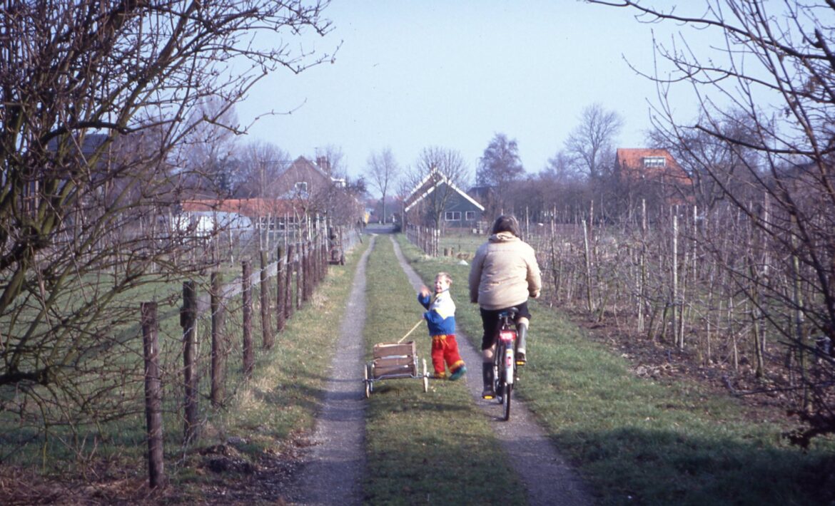 A picture from about 1990 where a small Jorik, dressed in bright colours, waves to the camera, as his grandmother cycles slowly back towards the farmhouse.