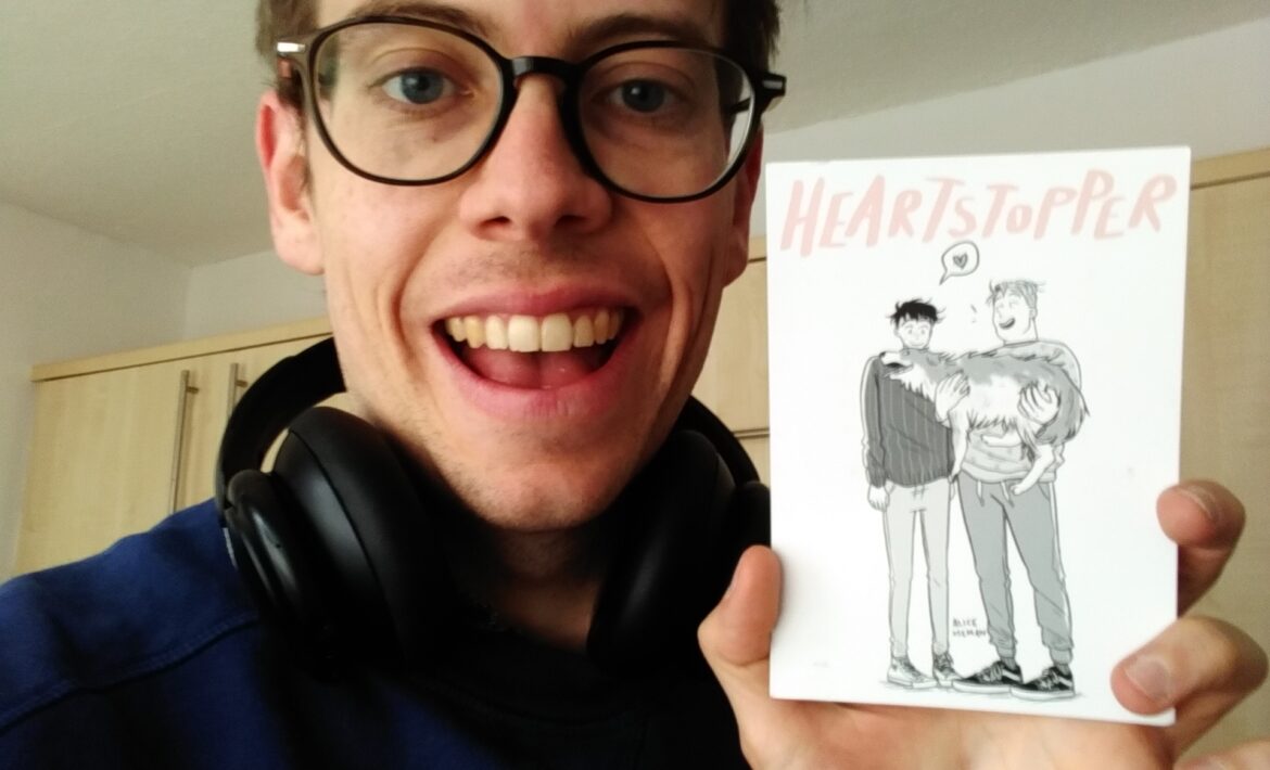 An image of Jorik, a 34-year old man, smiling a tired smile, holding up a postcard of Alice Oseman's Heartstopper, sent by his mother.